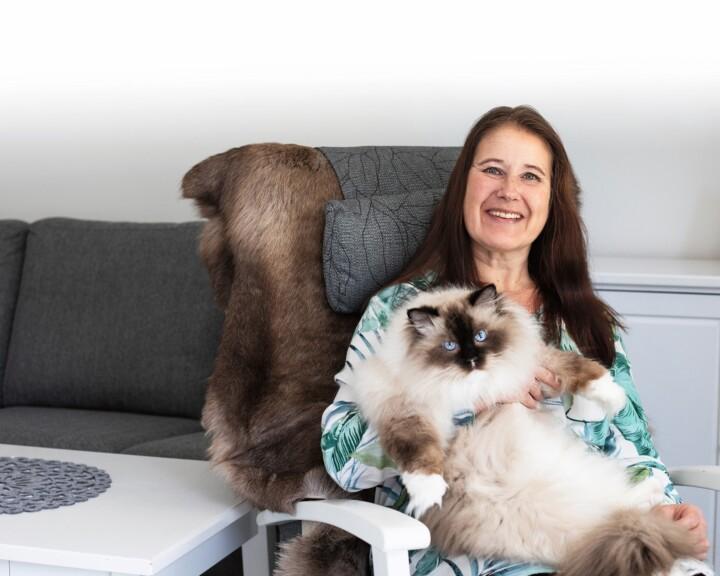 Kuvassa Henna Kastikainen istuu nojatuolissa kissa sylissään, katsoo kameraan ja hymyilee